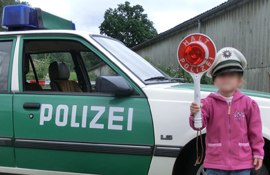 Kind mit Polizeikelle und -mütze vor einem Streifenwagen